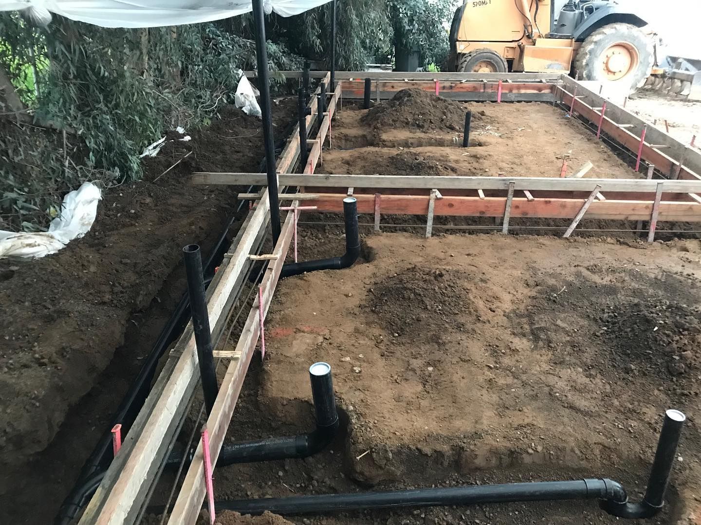 Construction site with black plumbing pipes in dirt trenches within wooden forms, ready for concrete.