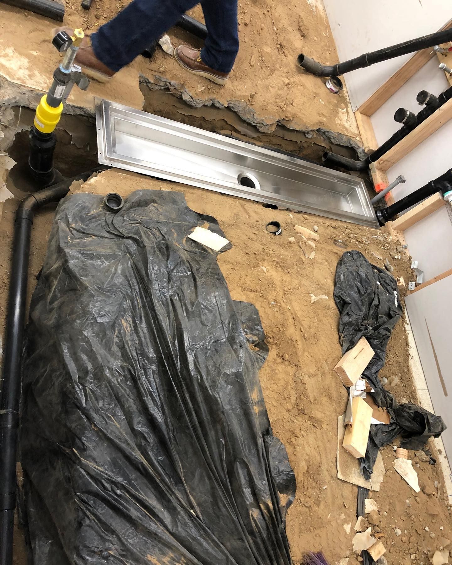 Plumbing work: Installation of a long, rectangular drain in a trench in a bathroom floor.  Black pipes, person in jeans.