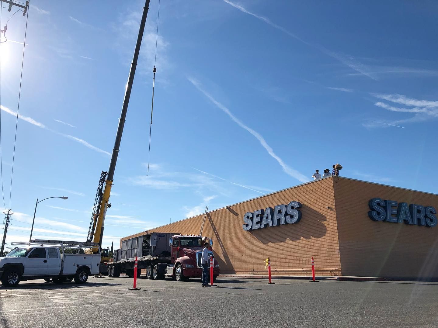 Crane lifting equipment near a Sears store under a blue sky. A truck and workers are present.