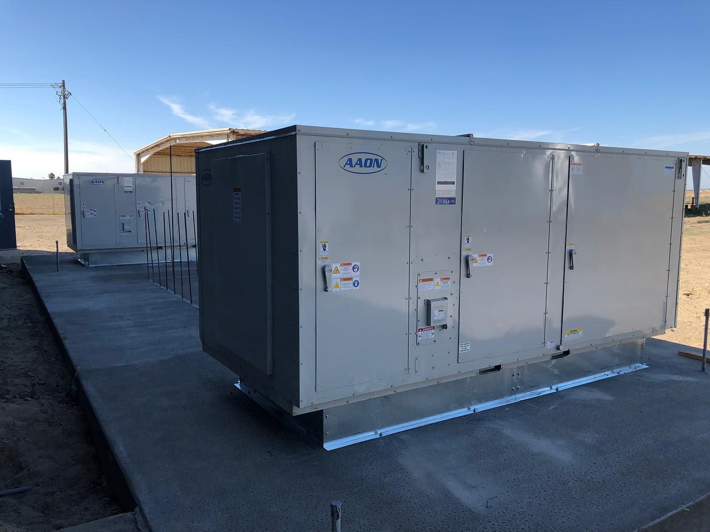 Two large, gray industrial air conditioning units on a concrete pad, outdoors under a clear blue sky.
