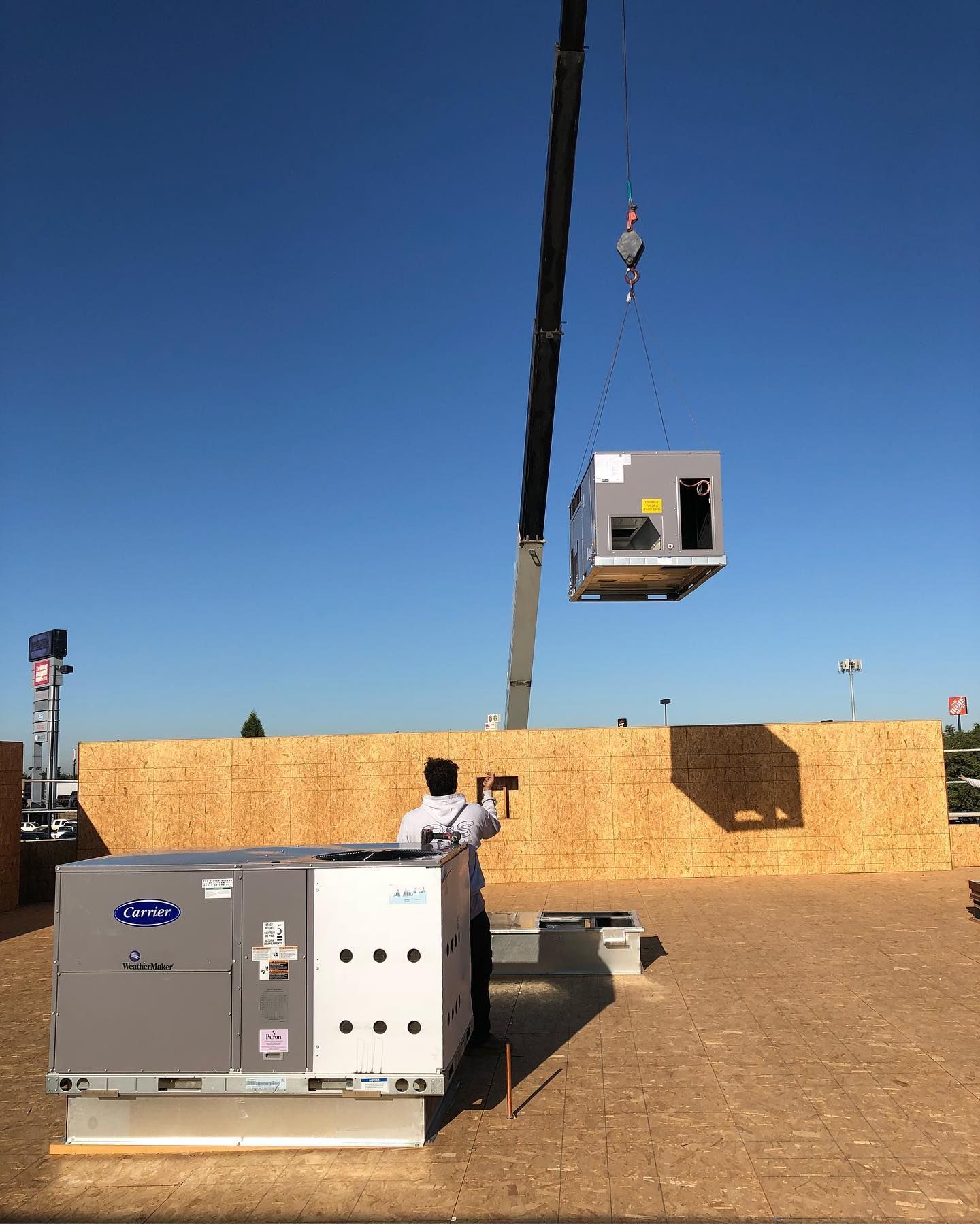Crane lifting an HVAC unit onto a rooftop. A worker stands nearby as another unit sits on the roof. Bright blue sky.