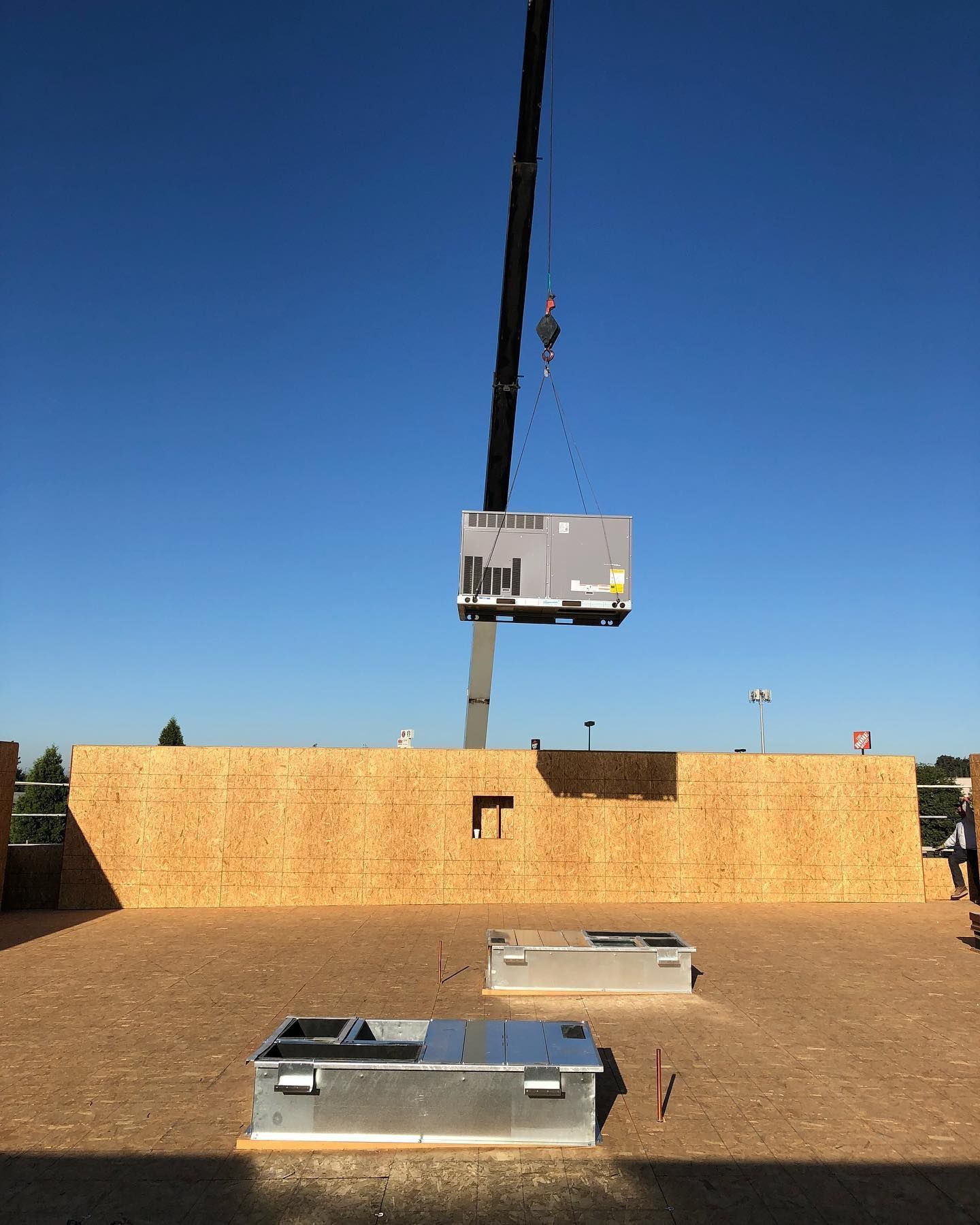 Crane lifting an HVAC unit above a construction site with plywood walls, blue sky.