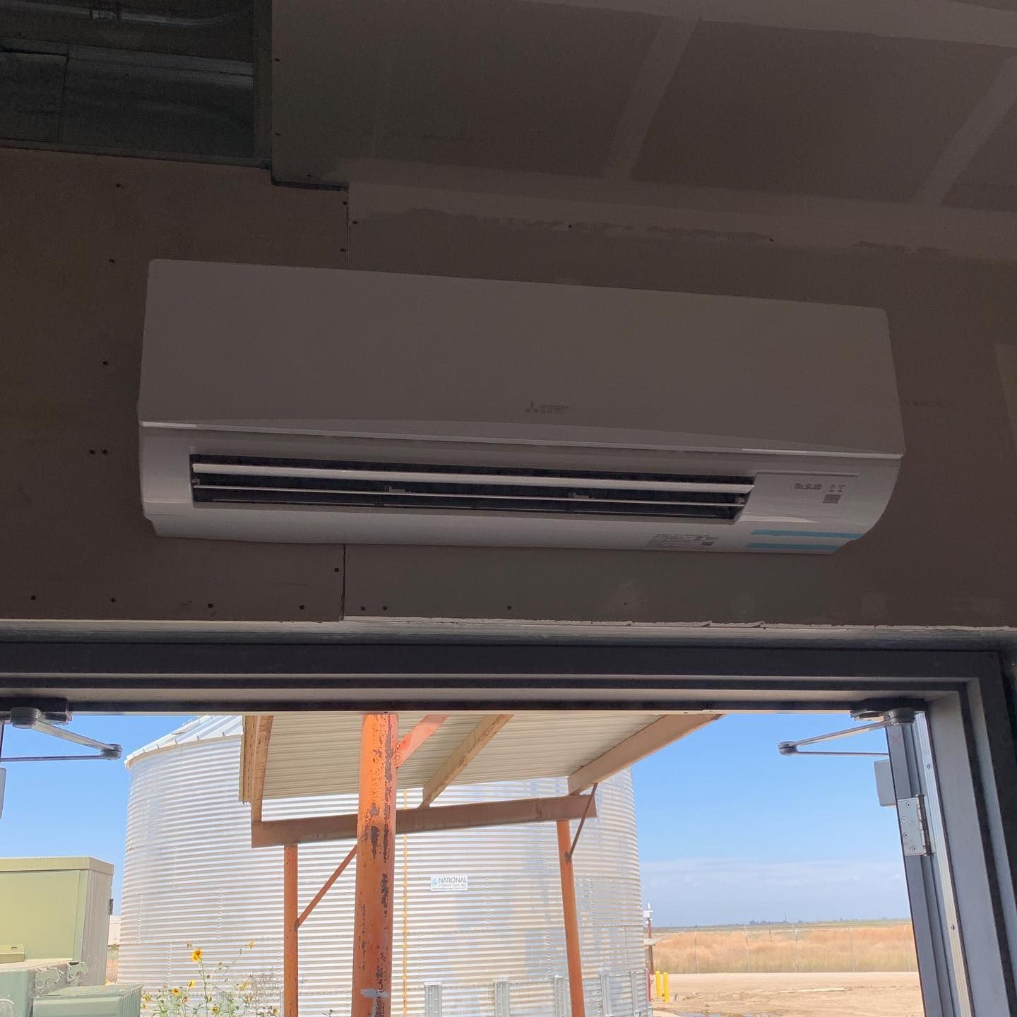 White air conditioner mounted above a door, with outdoor view of a silo and blue sky.