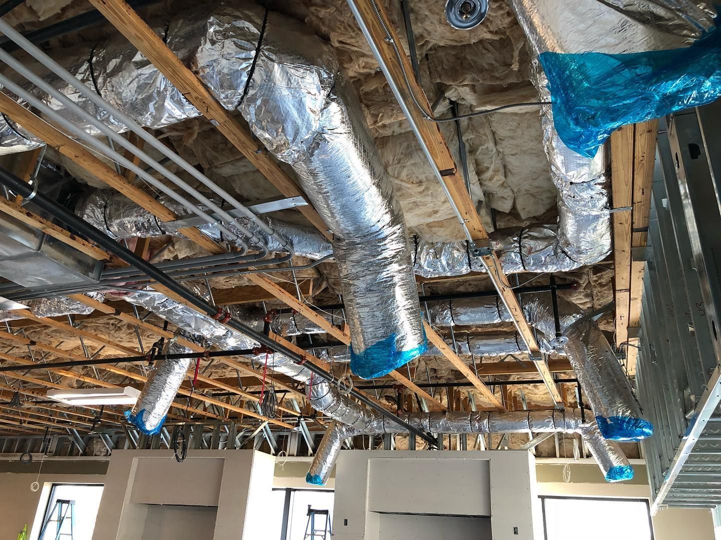 Construction site ceiling with ductwork wrapped in silver insulation and blue tape.