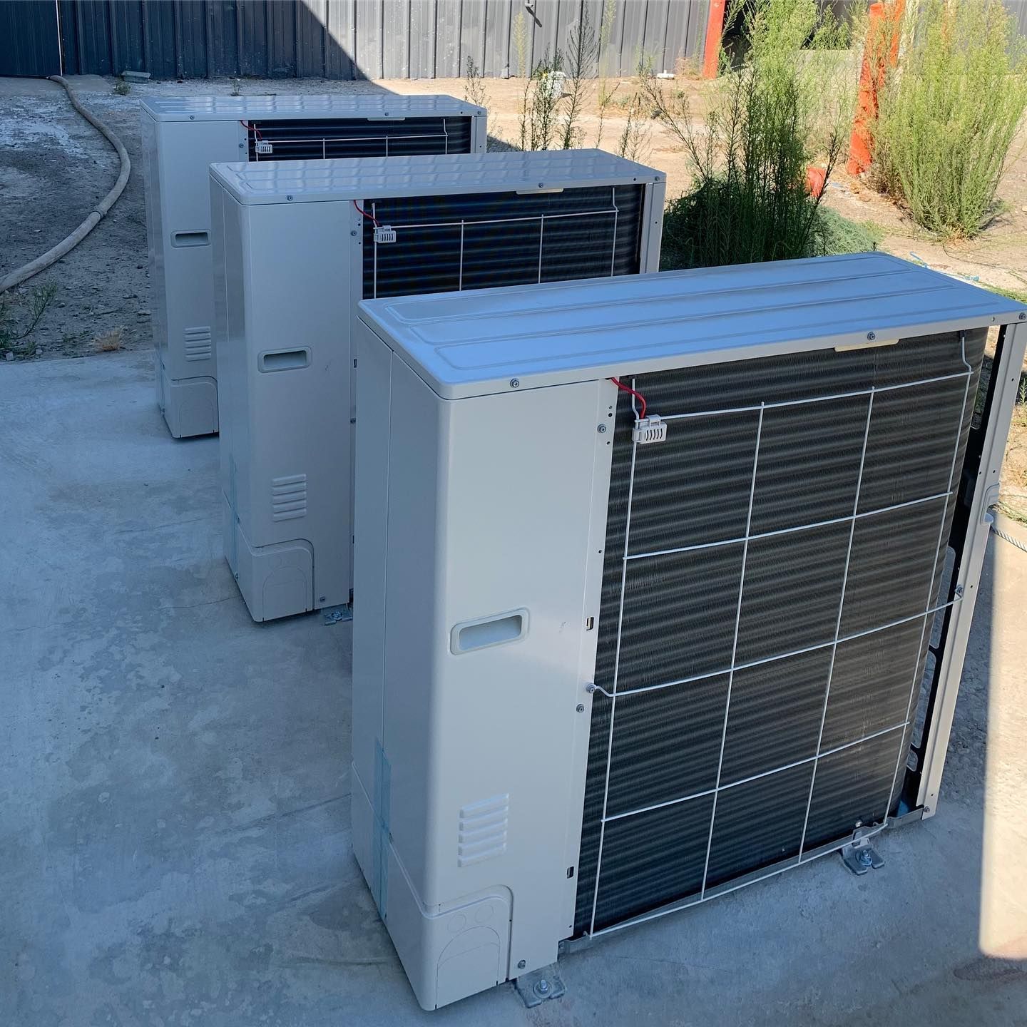 Three rectangular, off-white air conditioning units, lined up on concrete in front of a fence and some plants.