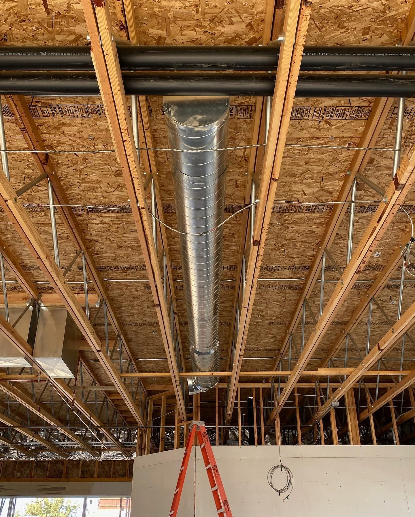 Construction site: ductwork and framing. Silver ventilation tube hangs from wood beams. Red ladder in foreground.
