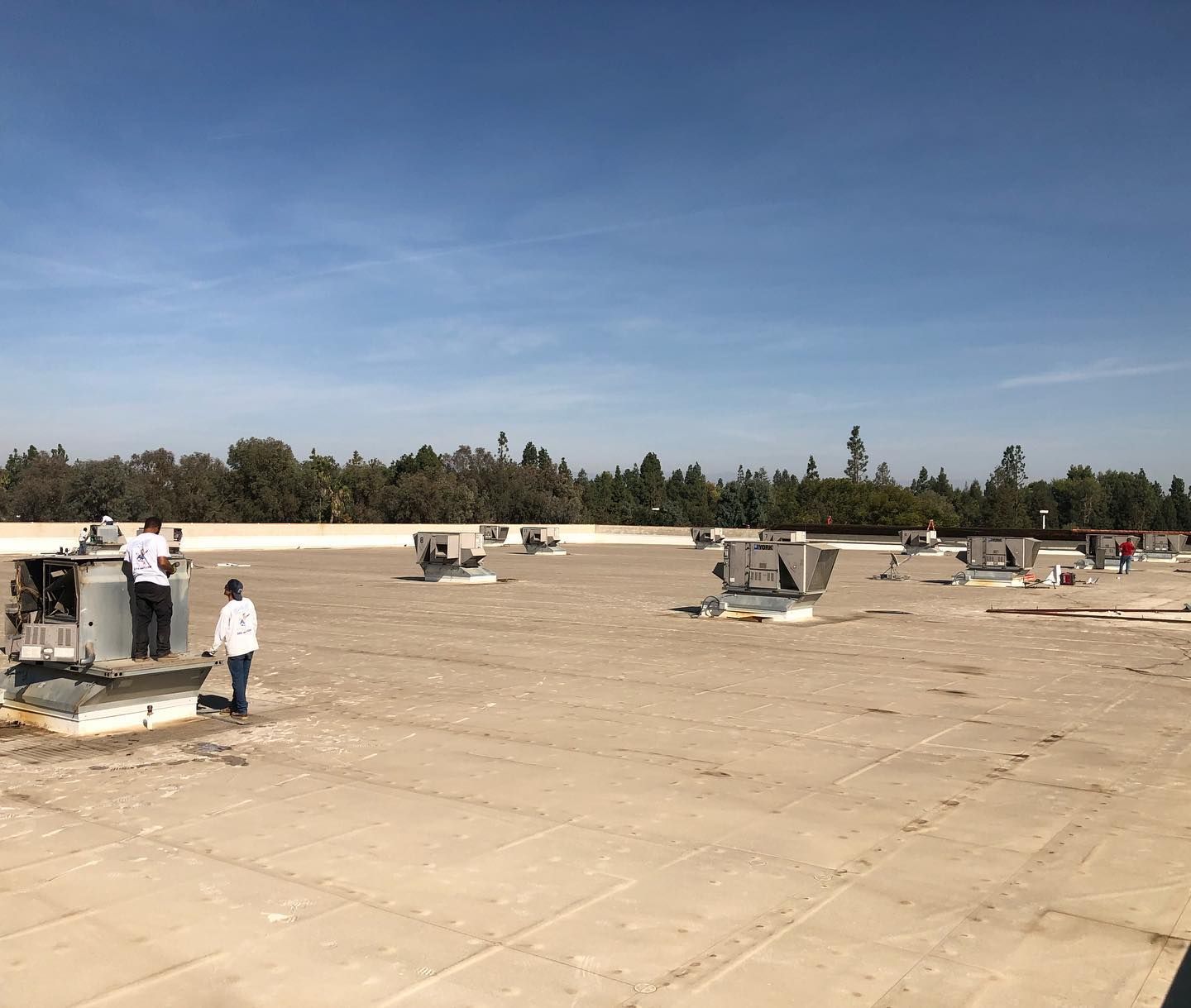 Workers on a flat roof with HVAC units. Blue sky, trees in the background, light-colored roofing material.