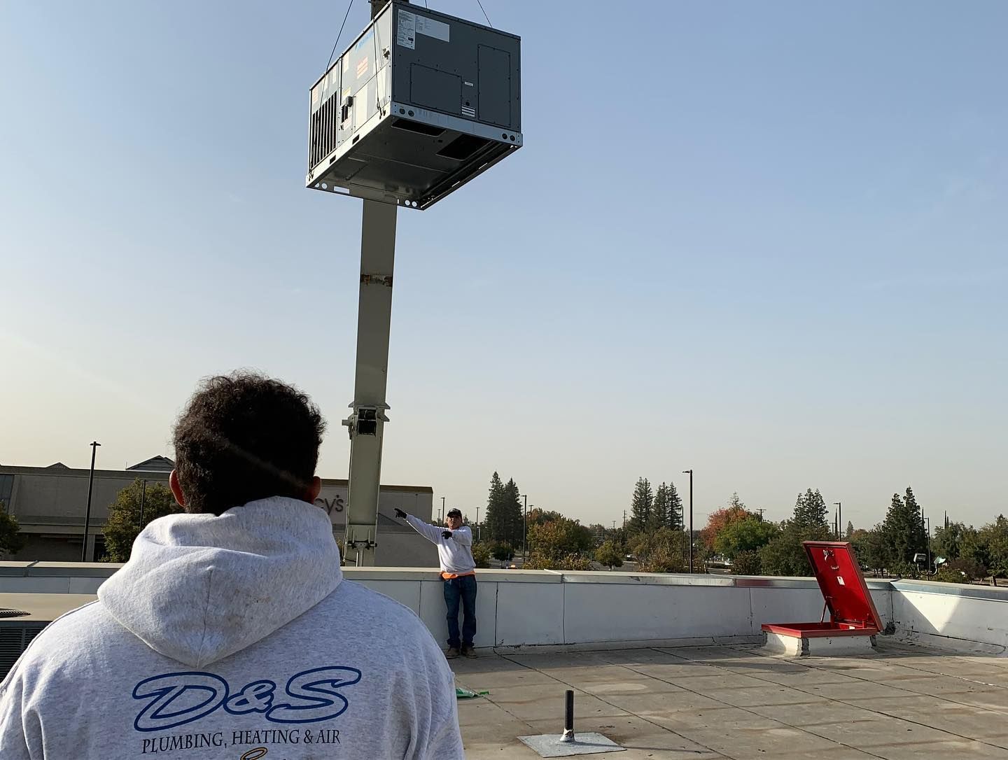 Workers installing HVAC unit on a roof. A crane lifts the unit, another worker directs it. Bright sky.