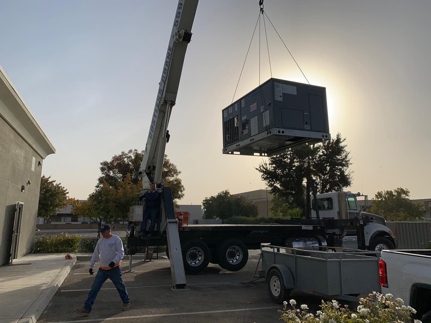 Crane lifting HVAC unit onto a truck. Man in work clothes watches. Bright sunlight.