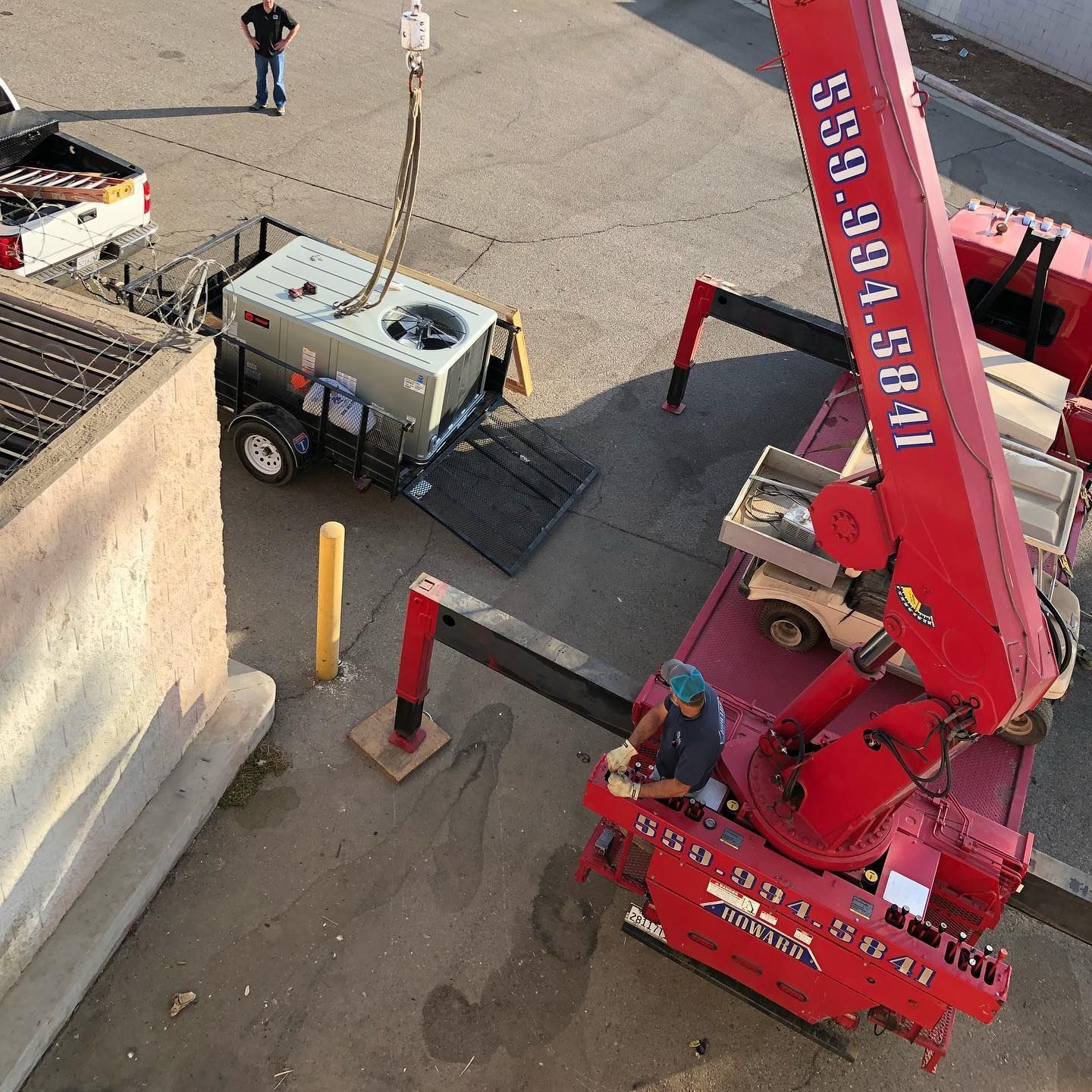 A crane lifts an HVAC unit off a trailer. Two people work, one in the crane. Outdoors, concrete.