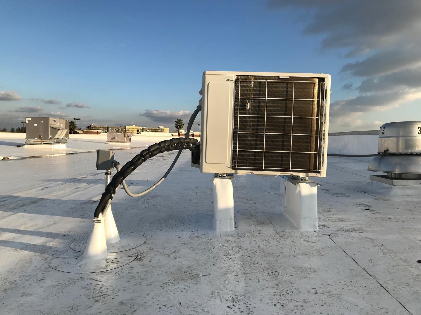 An air conditioning unit on a white rooftop, connected by black tubing. Blue sky background.