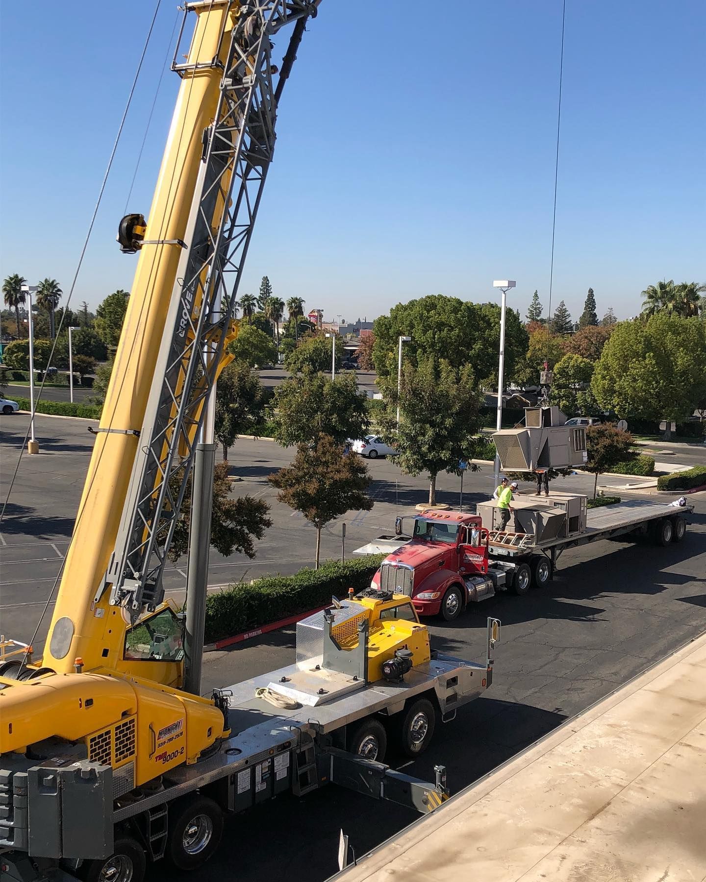 A large yellow crane lifting construction materials from a flatbed truck on a sunny day.