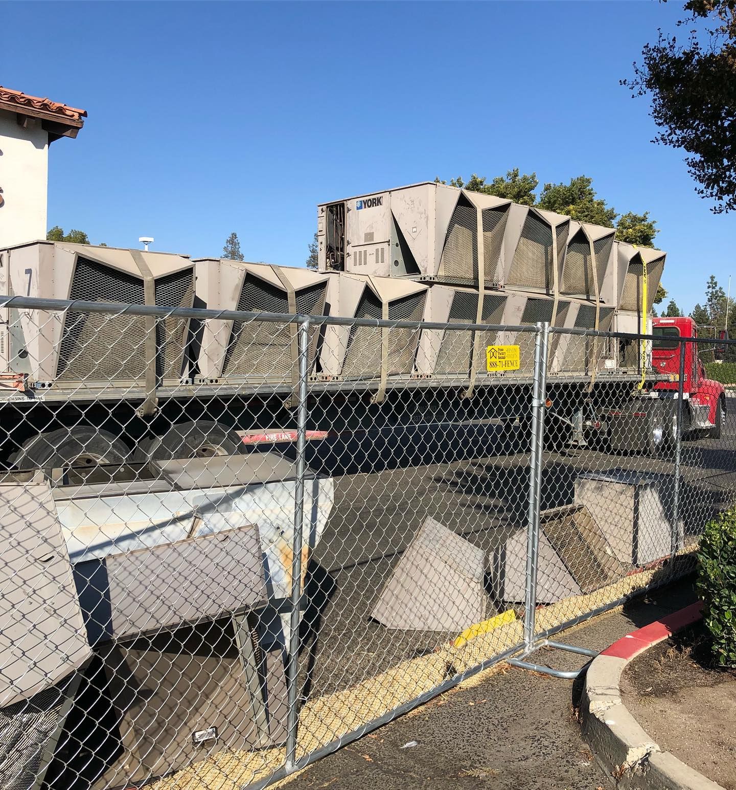 A flatbed truck carrying several large, gray HVAC units behind a chain-link fence.