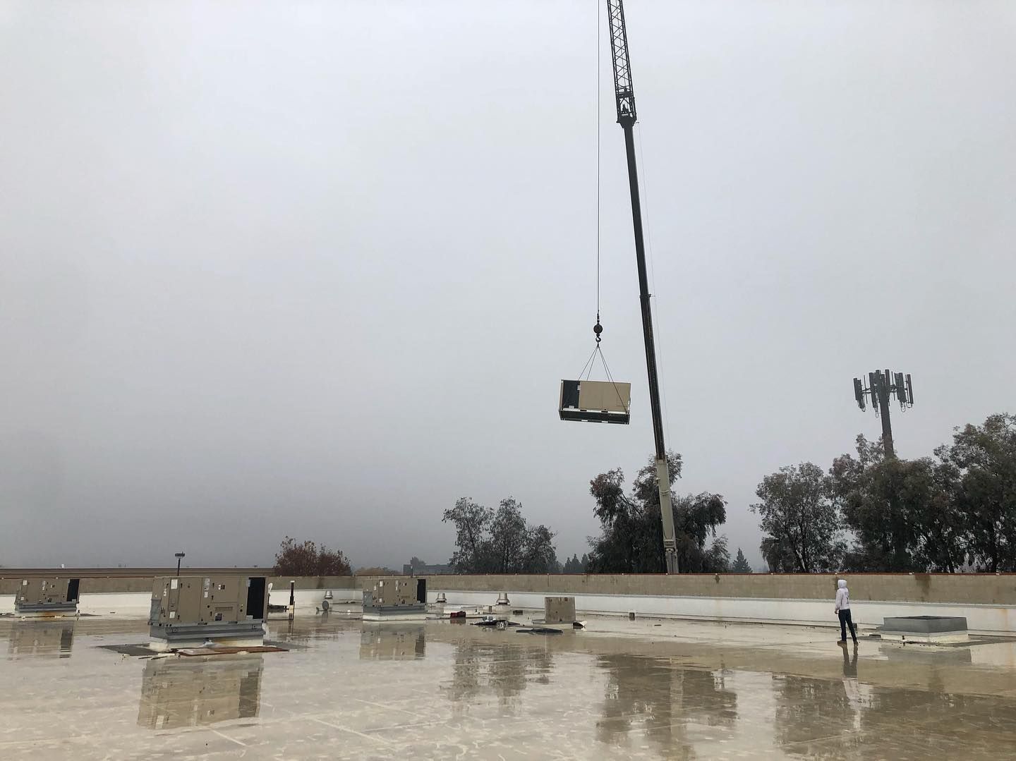 Crane lifting equipment onto a flat, wet roof; a worker stands nearby.