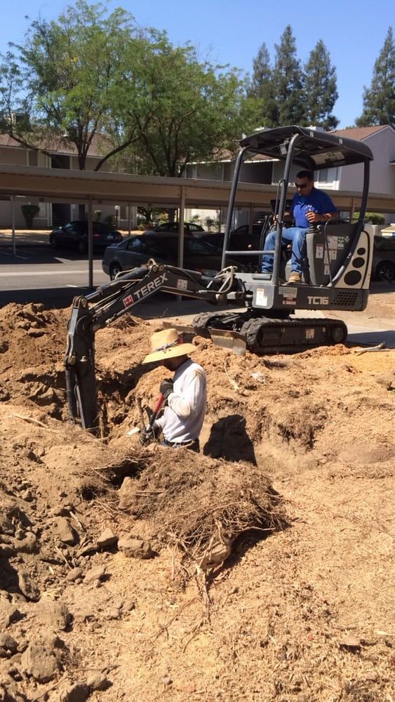 Man operates a small excavator while another works in a hole. Sunny day, residential setting.