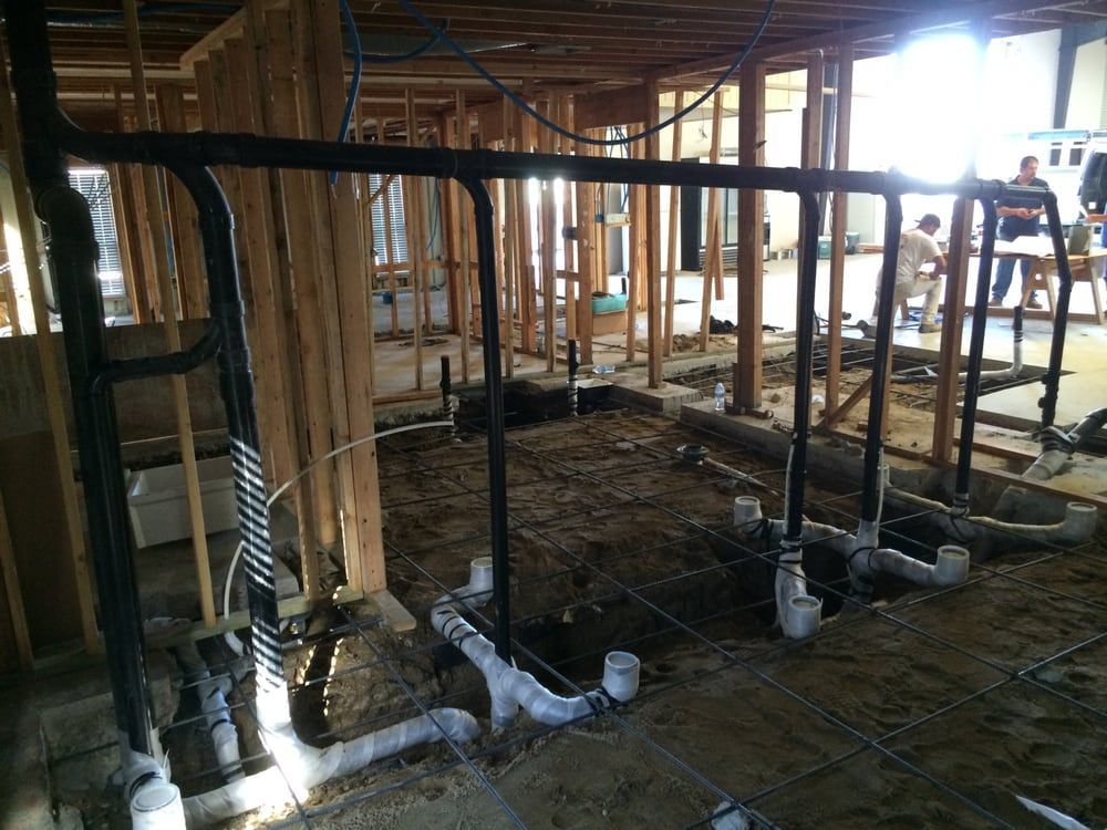 Interior view of a building under construction, showing plumbing and wooden frame. Men are in the background.