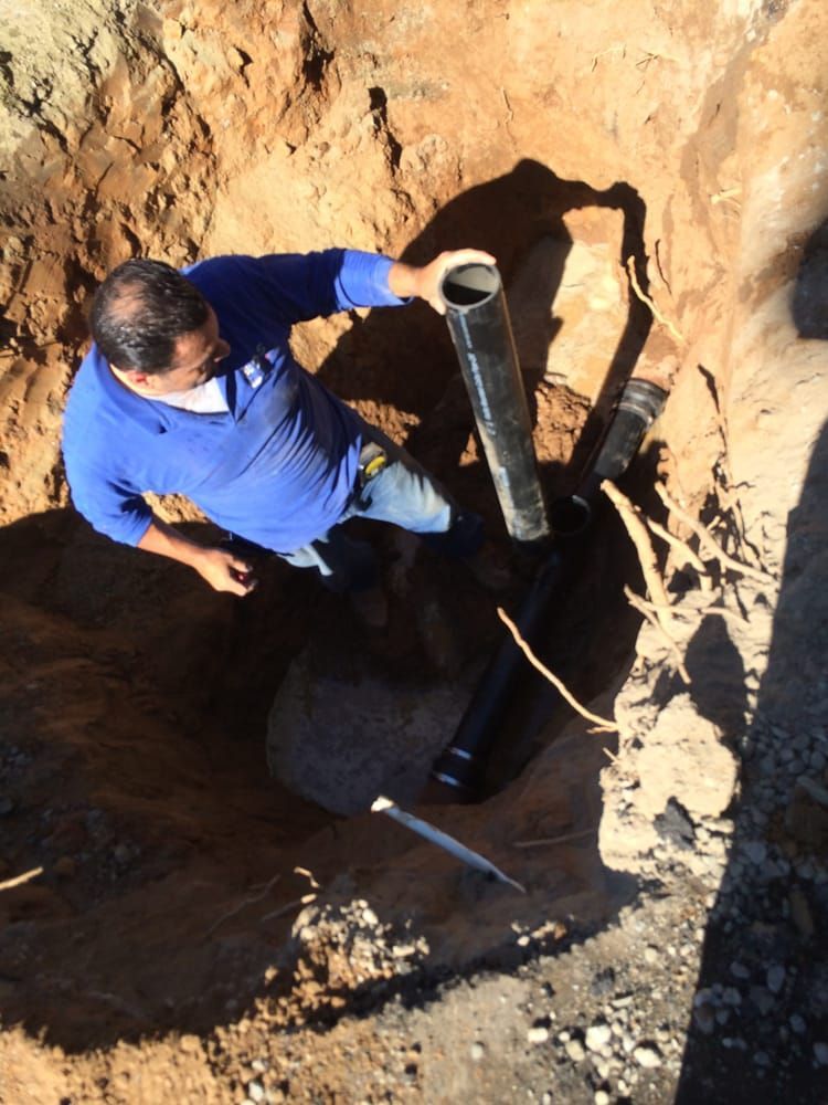 A person in a blue shirt holds a black pipe in an excavated trench.