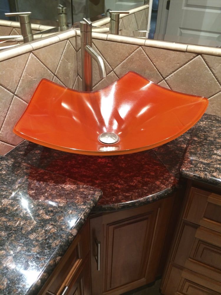 Orange glass vessel sink on granite countertop. Brown wooden cabinet below.