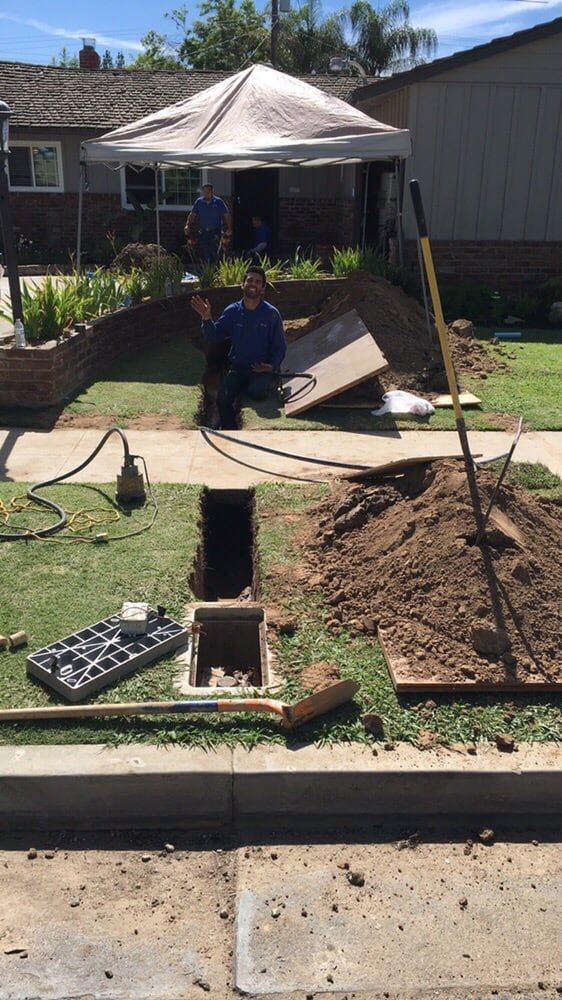 Workers are repairing something underground near a house. They are working in a hole next to a sidewalk.