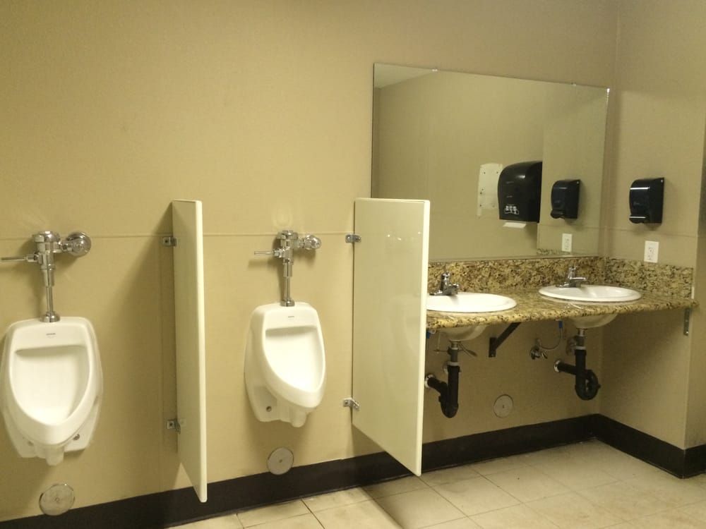 Public restroom with urinals, sinks, and a large mirror. Beige walls, light granite countertop.