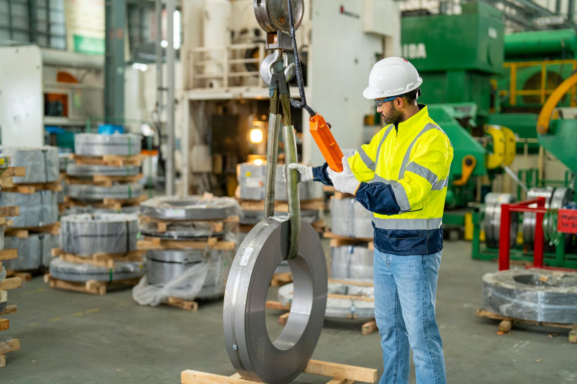 A male engineer, inside a metal manufacturing facility, inspects a crane with a remote controller.