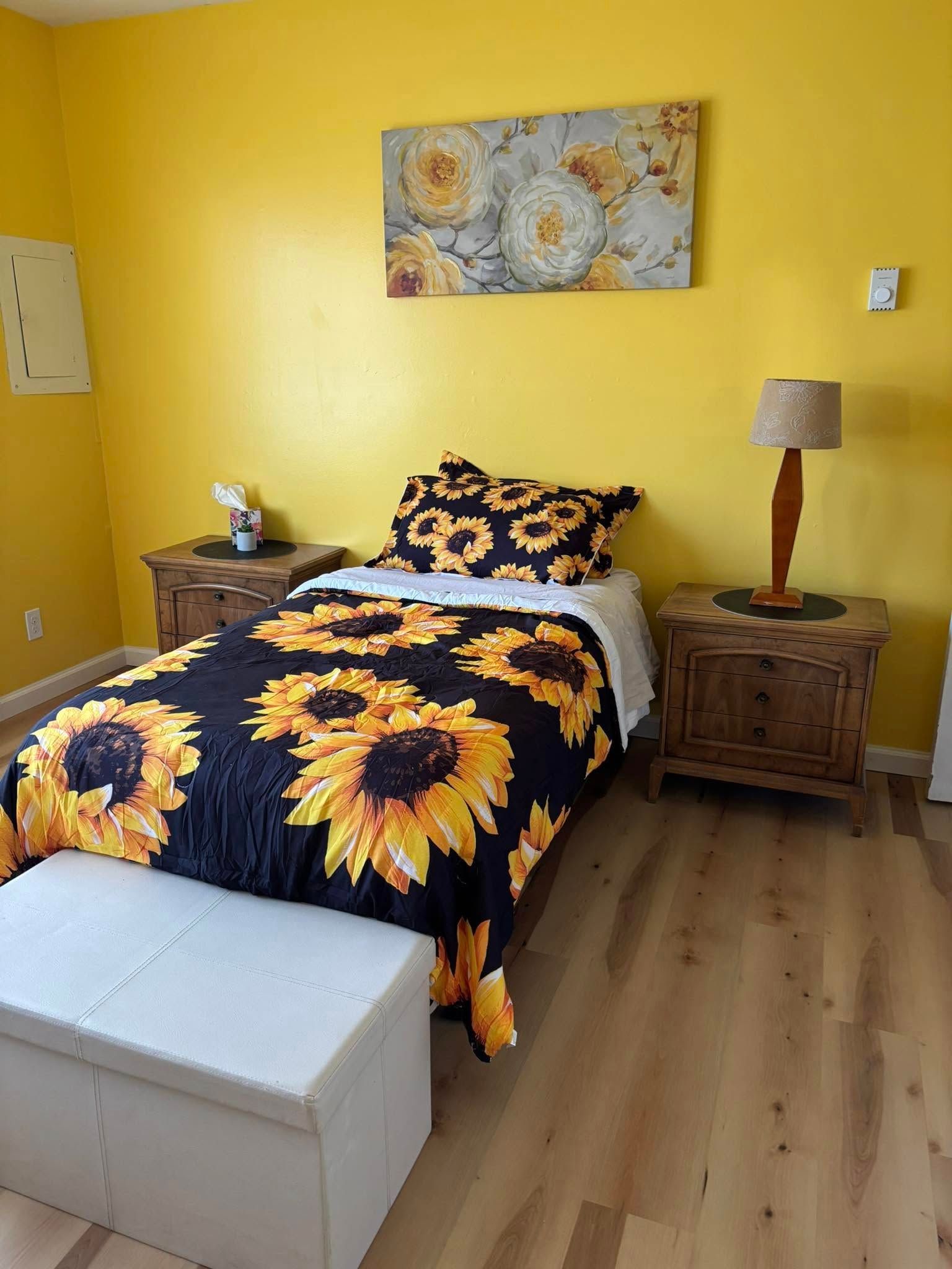 Yellow bedroom with a bed covered in sunflower bedding, wood bedside tables, and a white storage bench.