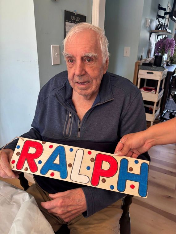 Elderly man, Ralph, holding wooden sign with his name, in a room. Sign has red, blue, and white letters.