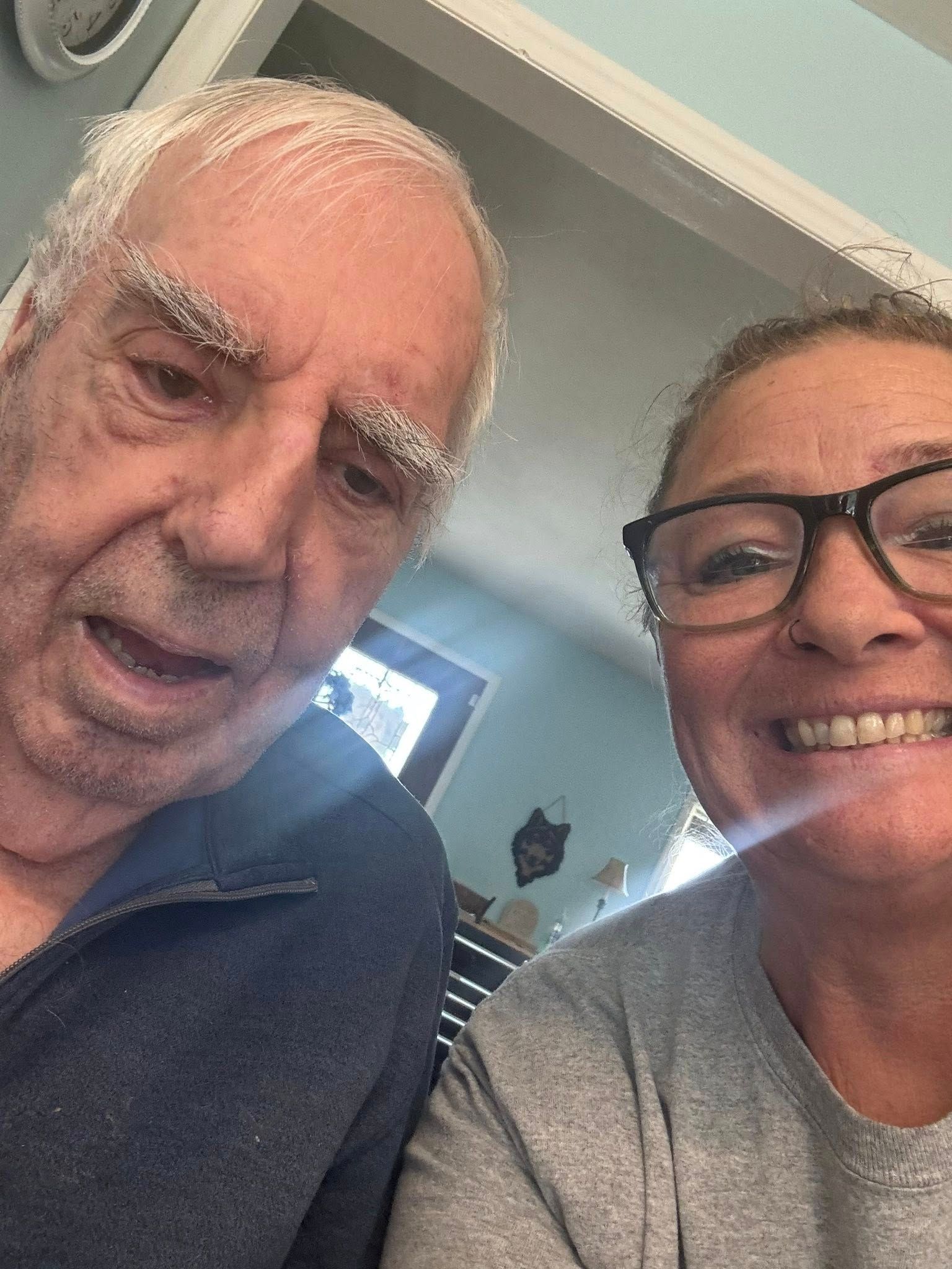 Man and woman smiling for selfie indoors; man's eyebrows prominent, woman wears glasses.