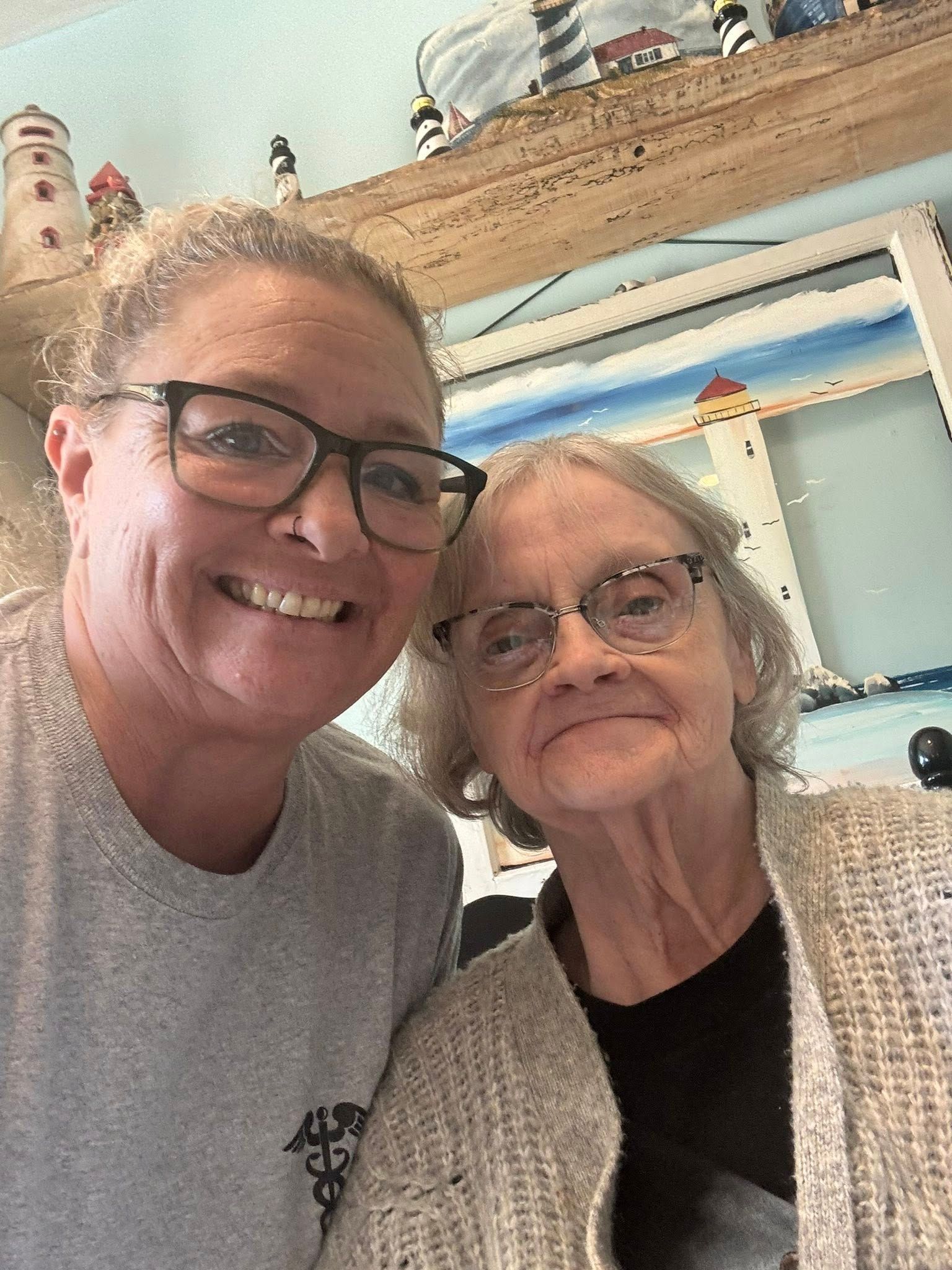 Two women smiling, close-up selfie. One with glasses, gray shirt. Other with glasses, cardigan. Nautical decor in background.