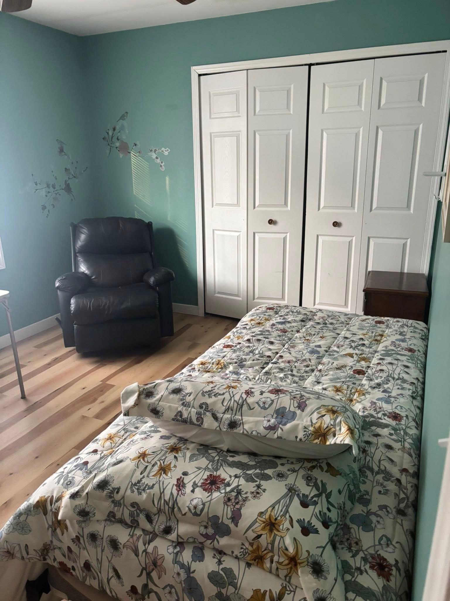 Bedroom with a floral bedspread, recliner, and white closet doors. Teal walls and light wood floors.