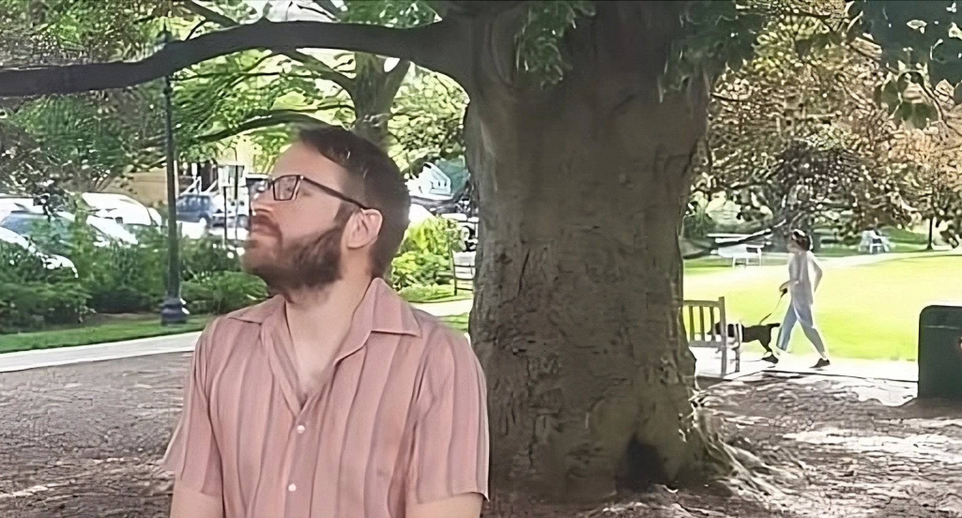 A man with glasses is standing next to a tree in a park.