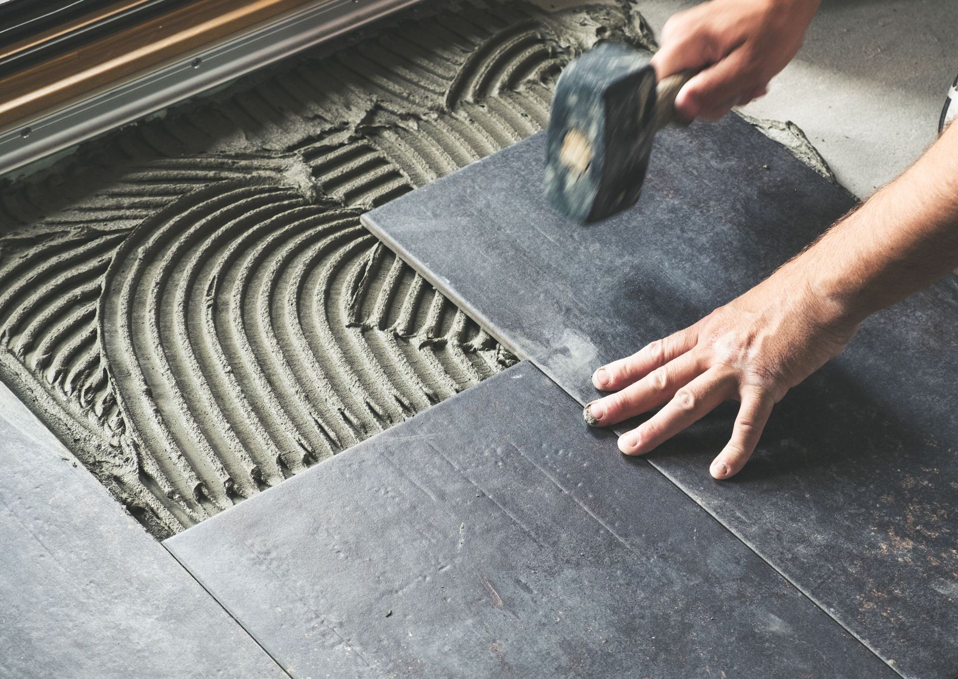 Person tiling a floor with a mallet, applying pressure to a dark gray tile over mortar.