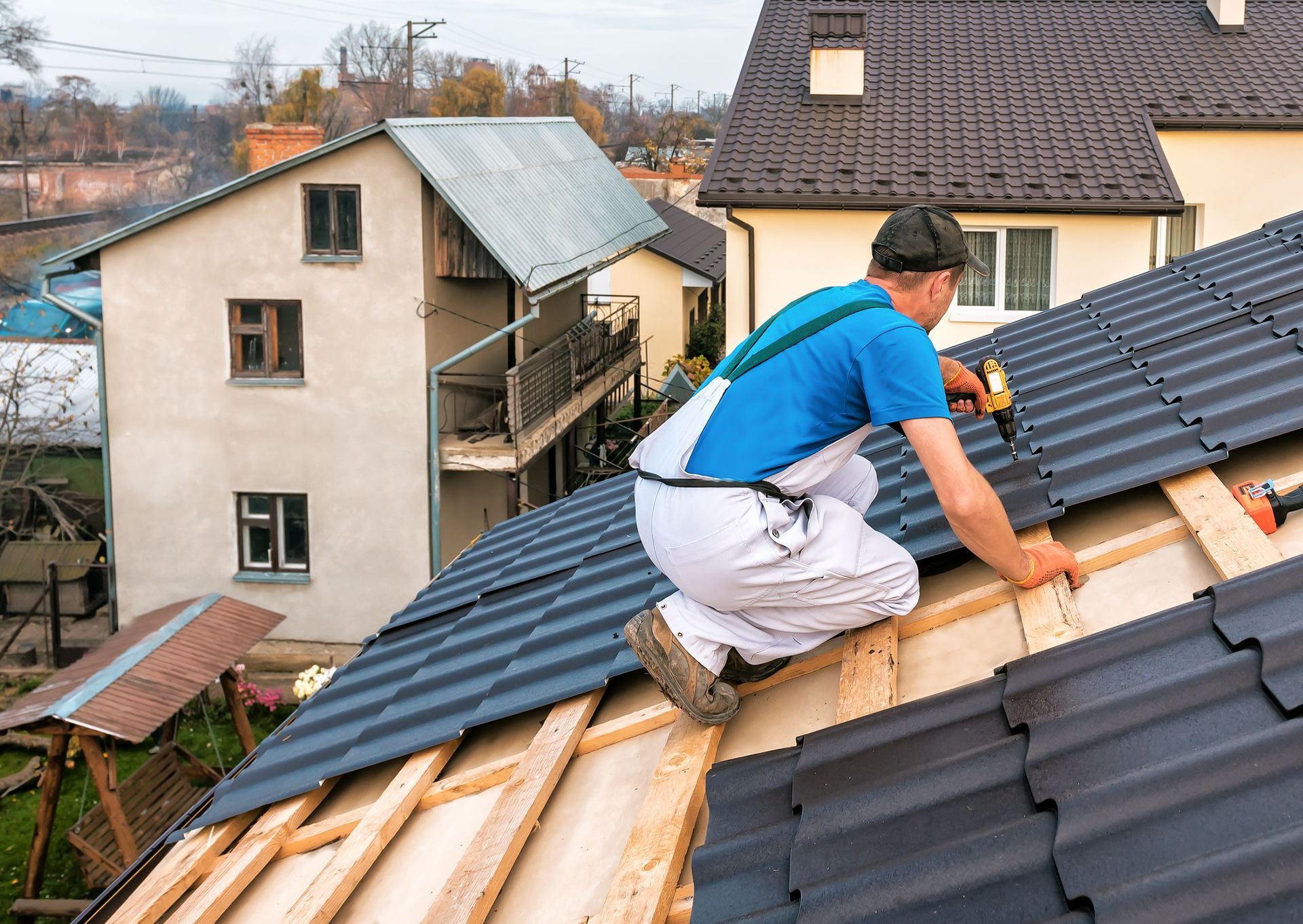 Roofer in blue shirt and white overalls on a rooftop, installing dark tiles.