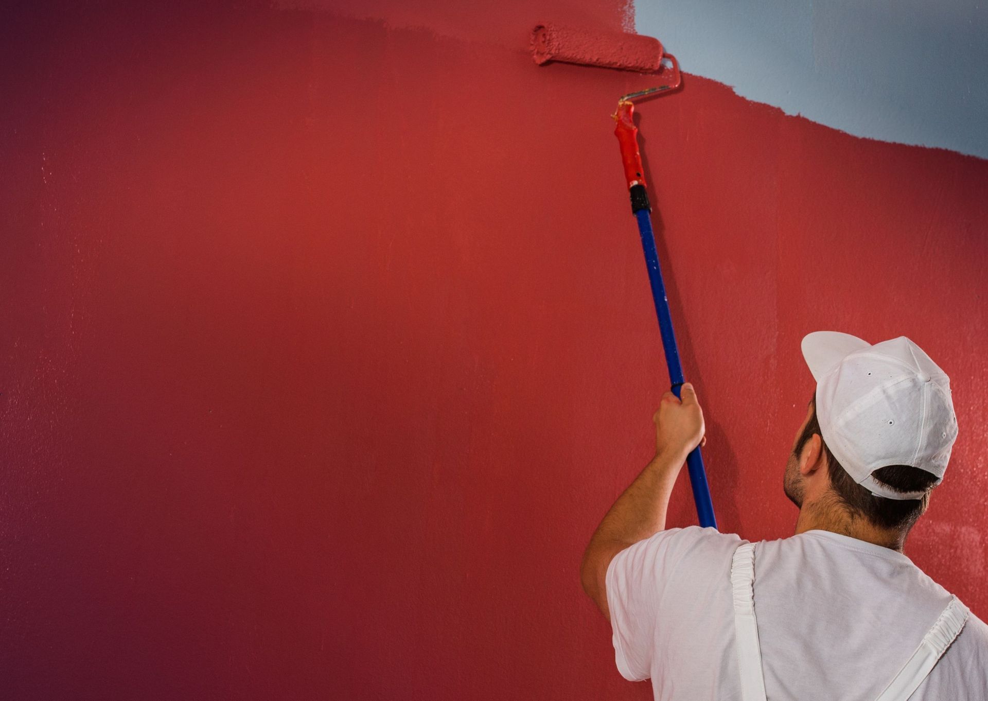 Person painting a red wall with a roller, wearing a white cap and shirt, against a blue ceiling.
