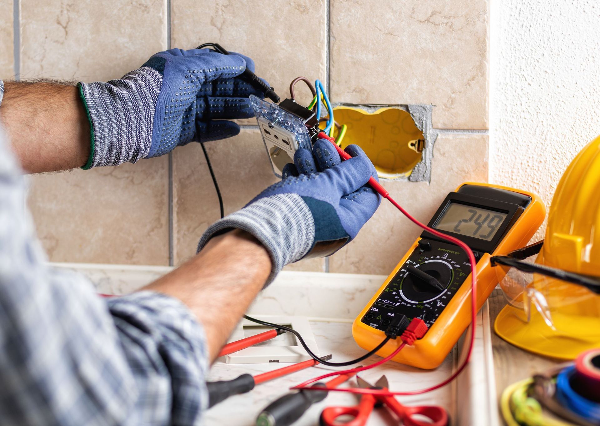 Electrician in gloves tests wires in wall outlet with a multimeter, beside tools and hardhat.