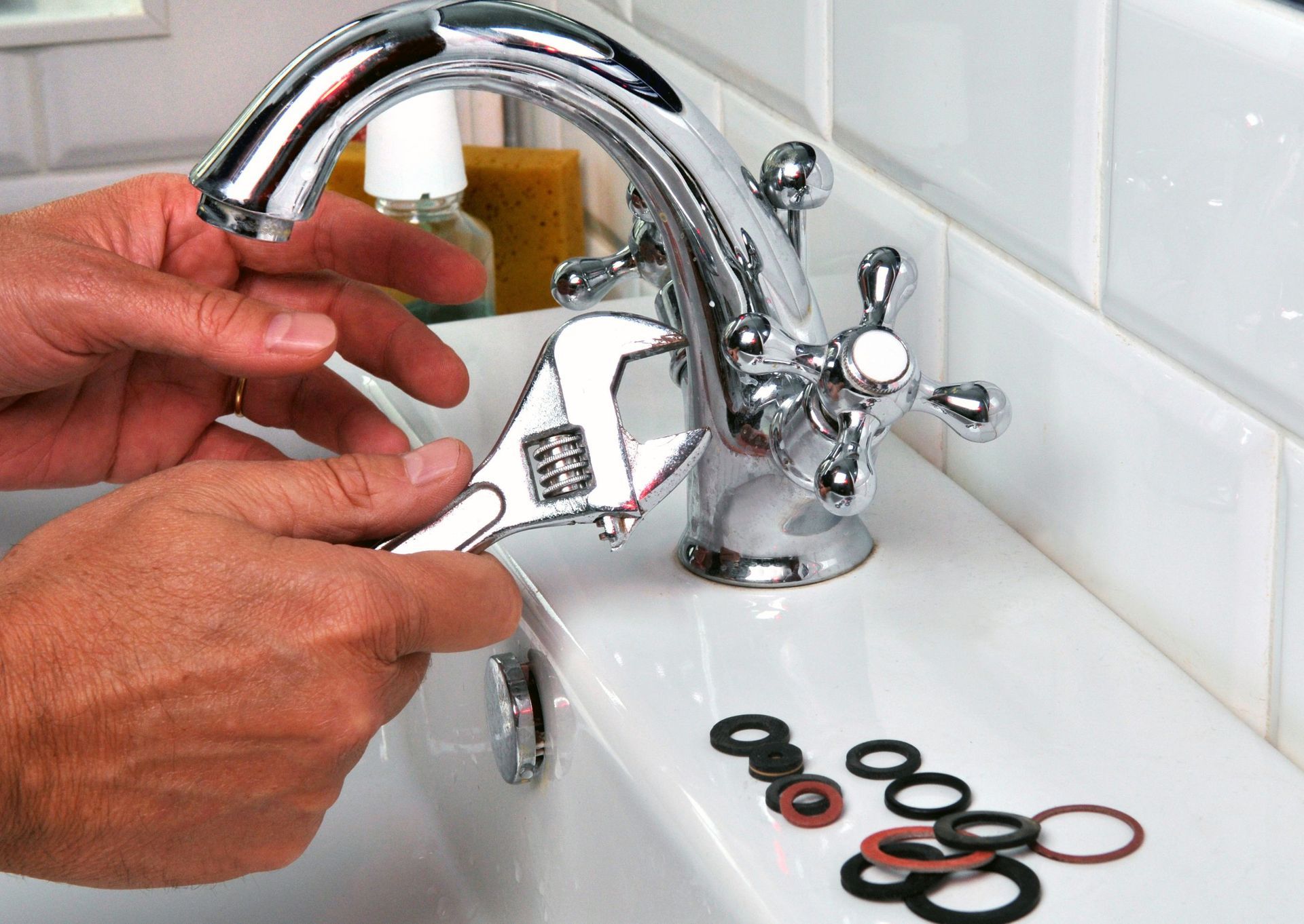 Hands using a wrench to repair a chrome faucet in a bathroom sink, various washers visible.
