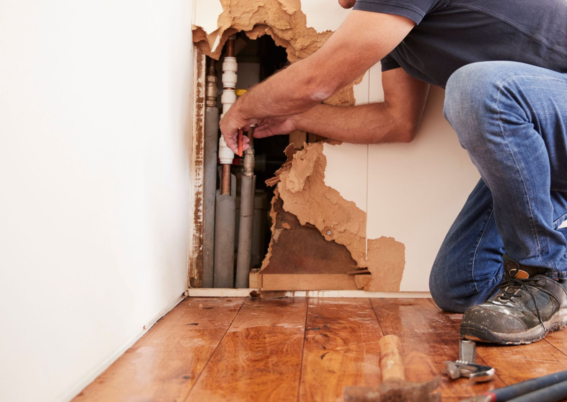 Man kneeling, fixing pipes in damaged wall, surrounded by wood flooring.