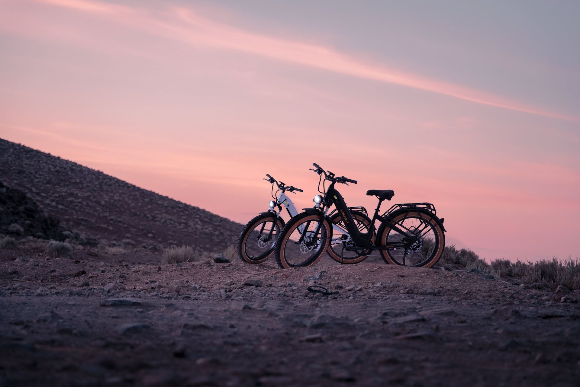 Two bicycles are parked on top of a dirt hill at sunset.