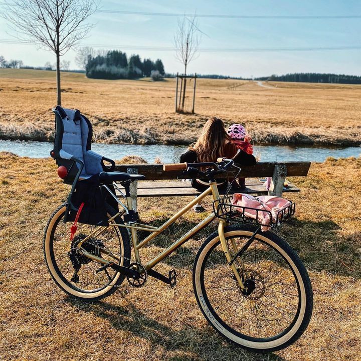 A woman sits on a bench next to a bicycle with a baby seat attached to it.