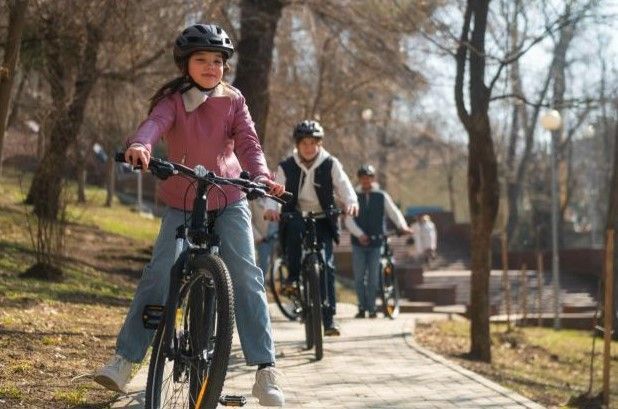 Girl in pink jacket rides bike in park with friends, all wearing helmets.