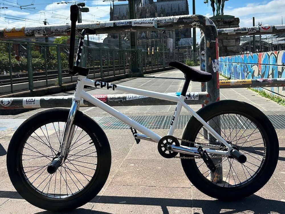 A white bmx bike is parked on the sidewalk next to a fence.