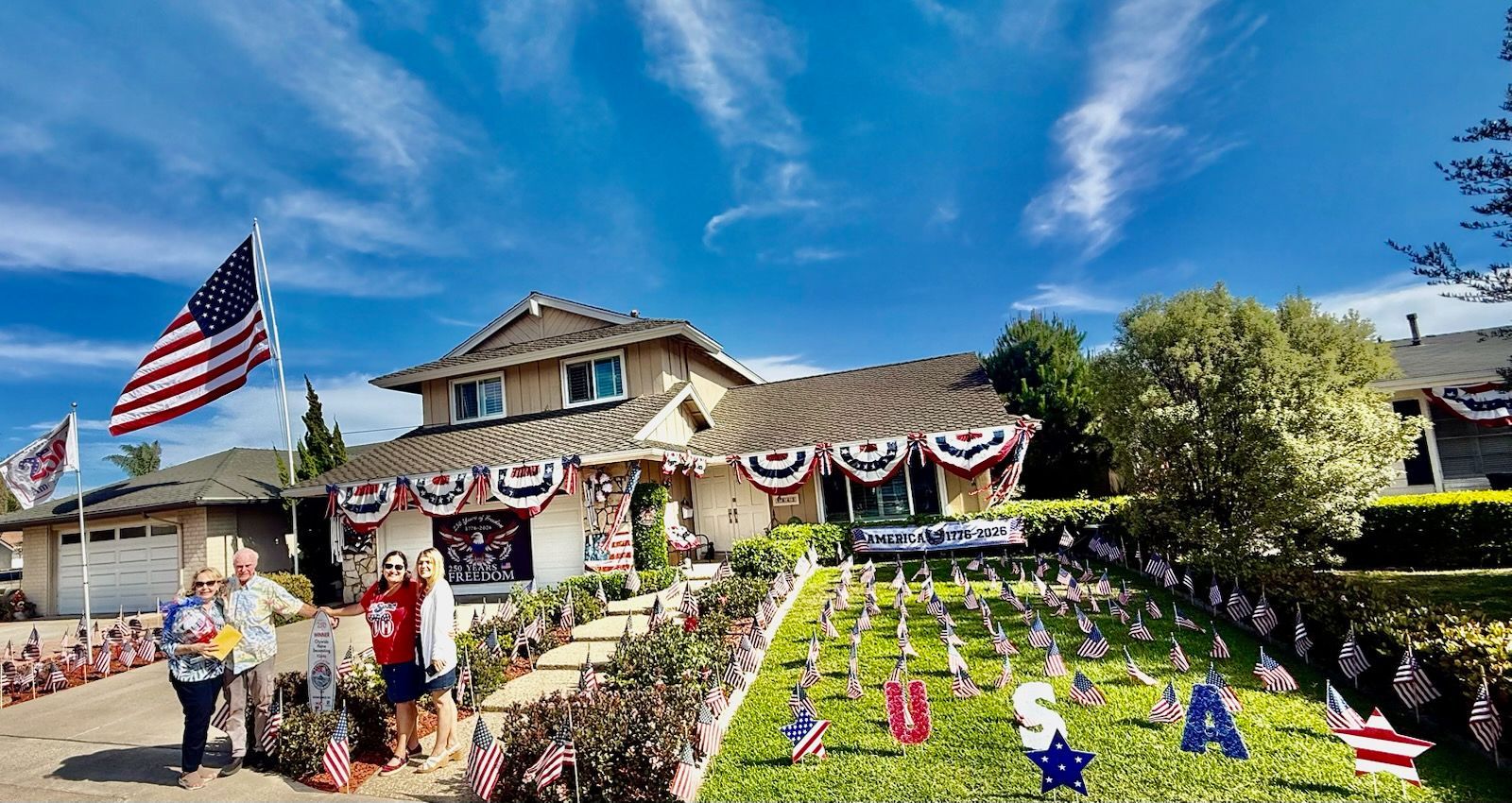 House decorated with patriotic bunting and U.S. flags, with red-white-blue lawn letters under a bright sky