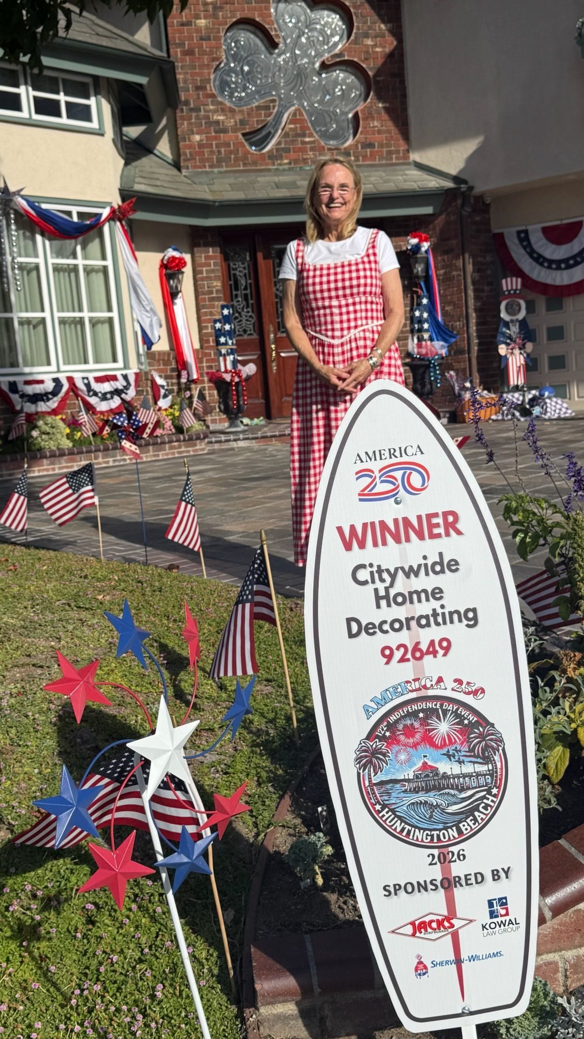 Woman in red-checkered dress beside a “Winner” sign on a decorated lawn with patriotic bunting and stars.