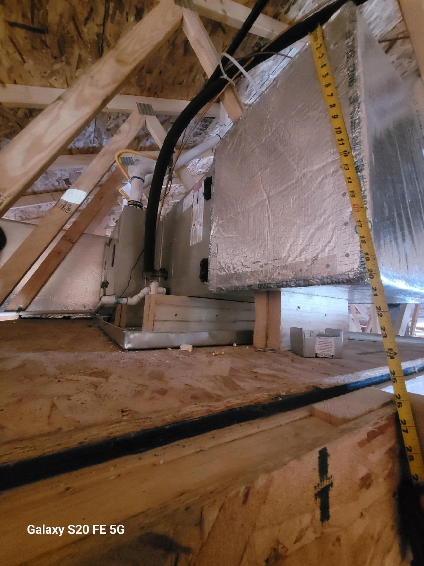 Attic view of an HVAC unit. Wooden rafters, ductwork, and measuring tape are visible.