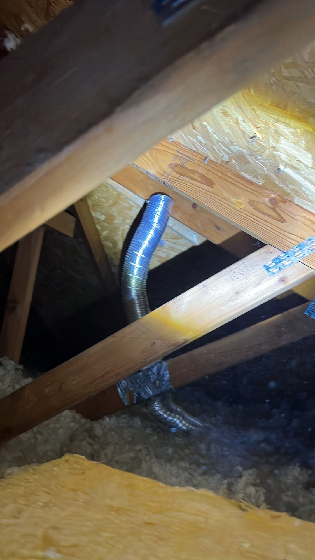Flexible metallic ducting in an attic space, running through wooden rafters, with insulation visible.