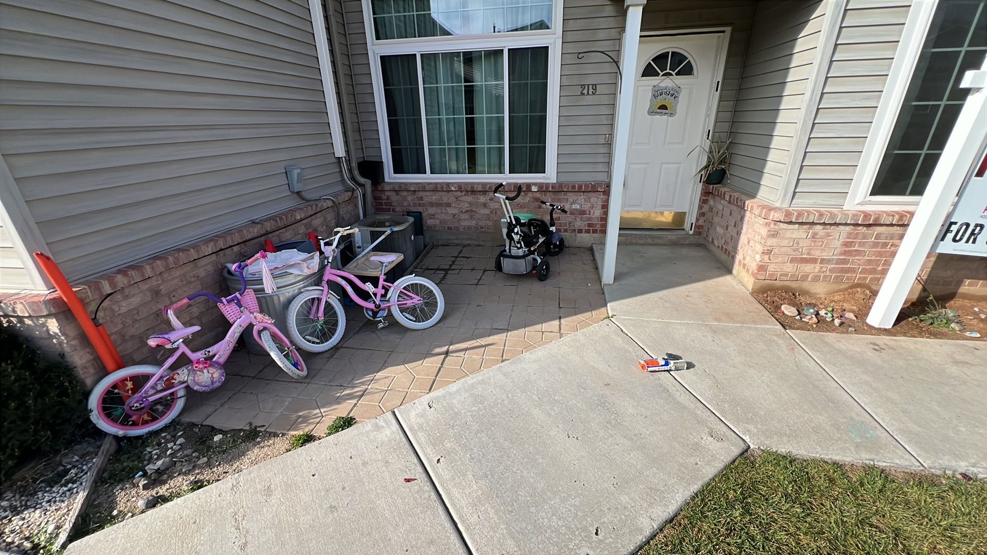 Pink bicycles, stroller, and pressure washer sit on a porch next to siding and a door.