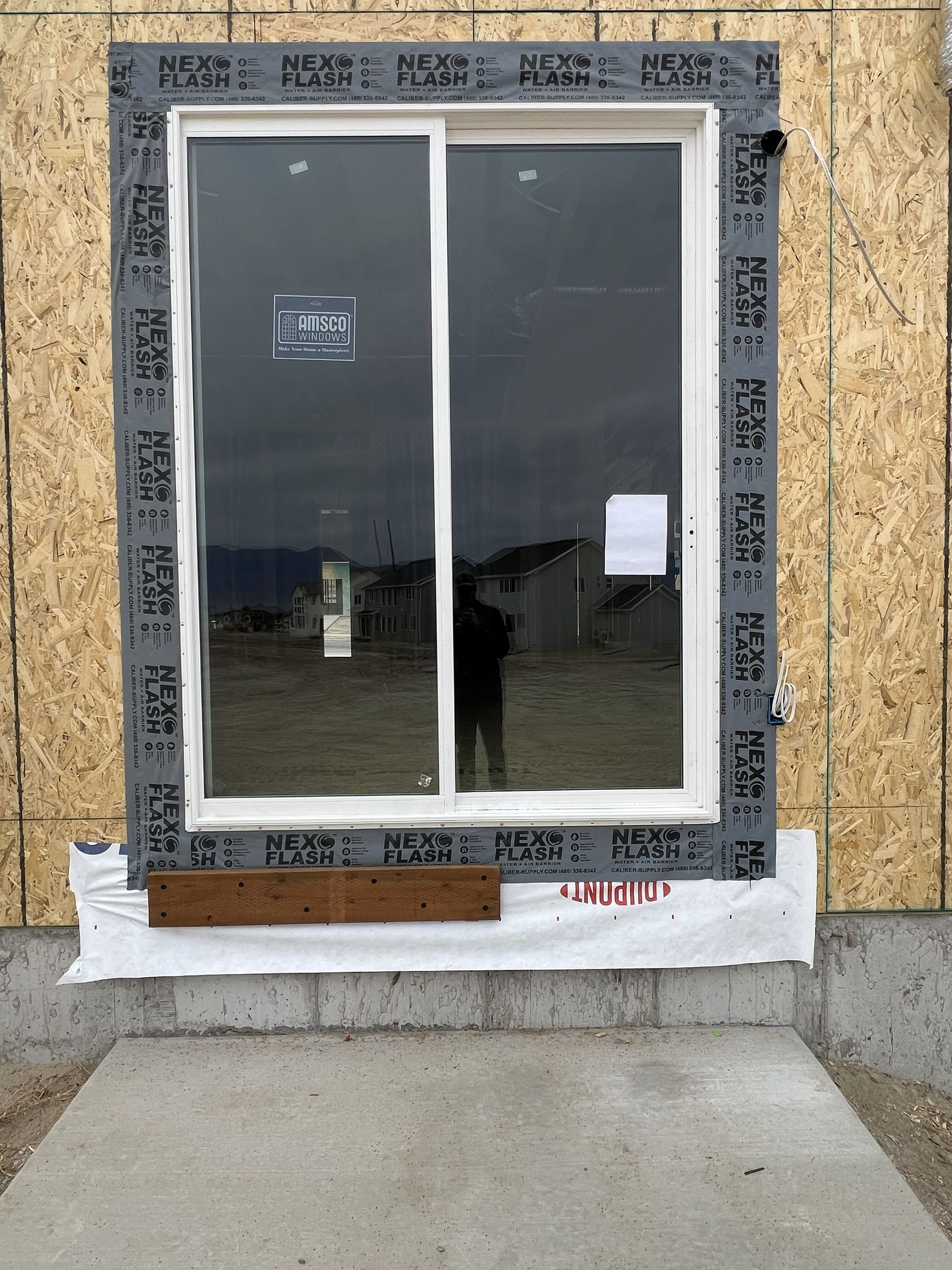 Window installation: Exterior view of a sliding glass window in a wooden frame with black flashing tape, over a concrete foundation.