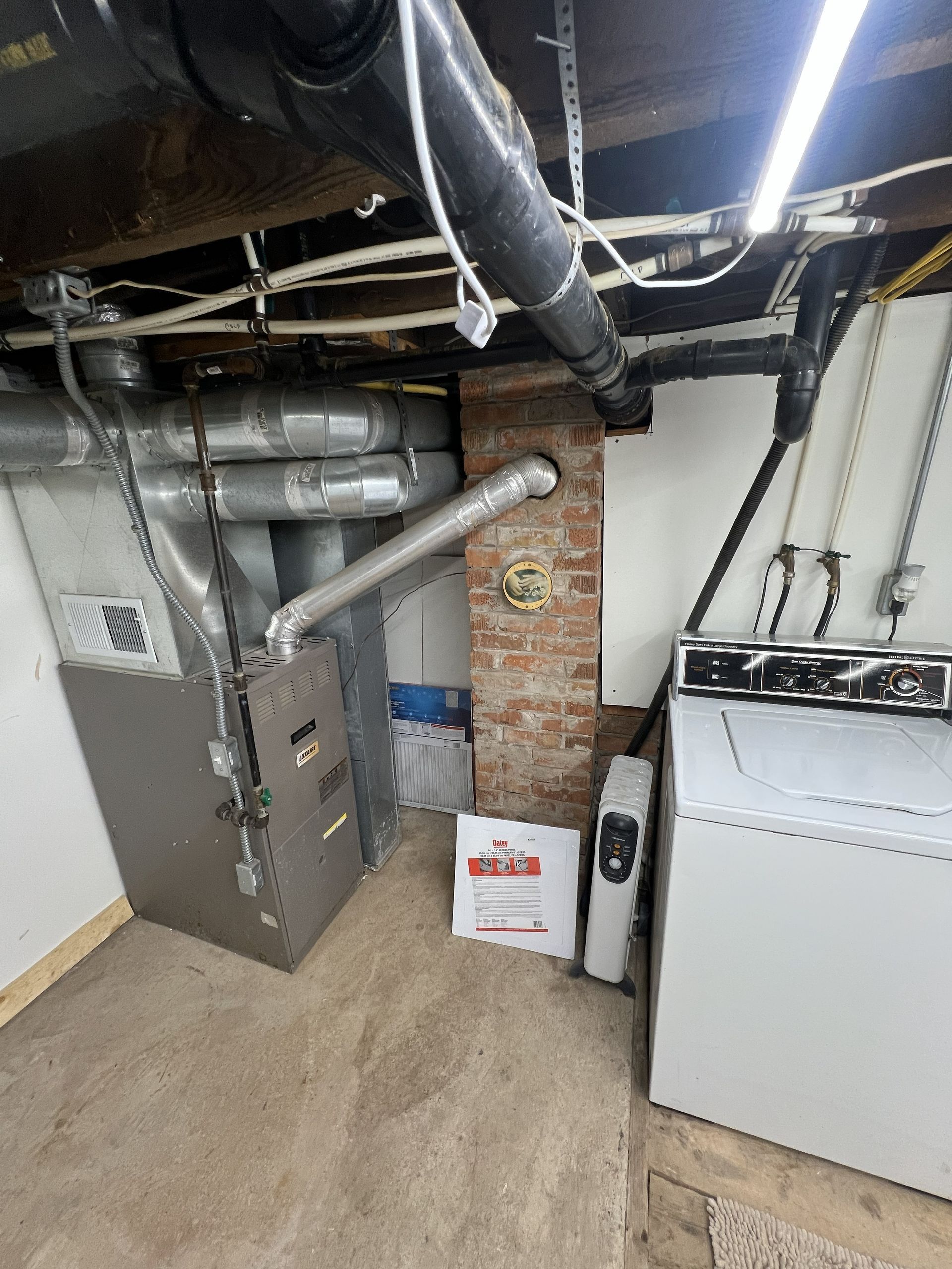 Furnace, brick pillar, and washing machine in a basement with exposed pipes and ductwork.