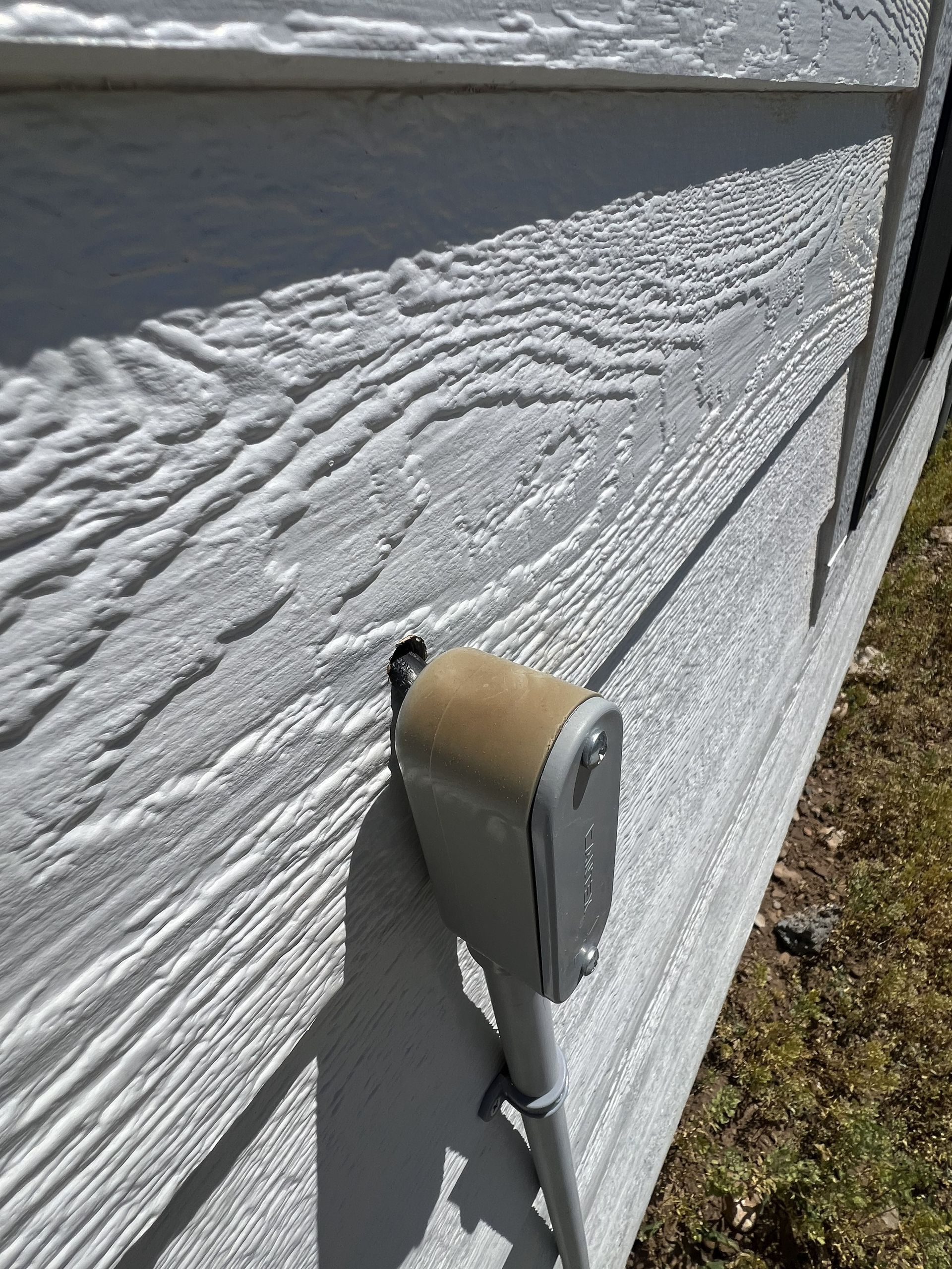 A beige electrical box attached to white wood-look siding, with a pipe running down.