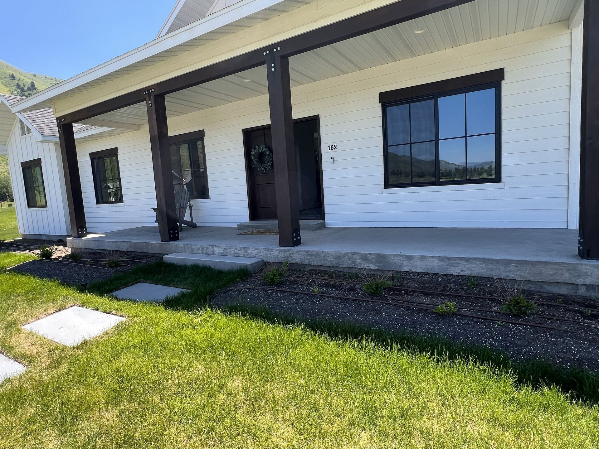 White farmhouse with dark trim and porch, green lawn, mountains in the background.